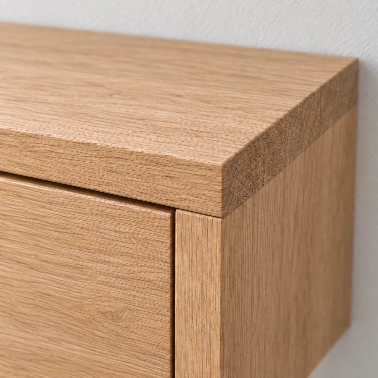 Close-up of a light oak wood cabinet corner showing the drawer front, top surface, and wood grain texture against a plain light wall.