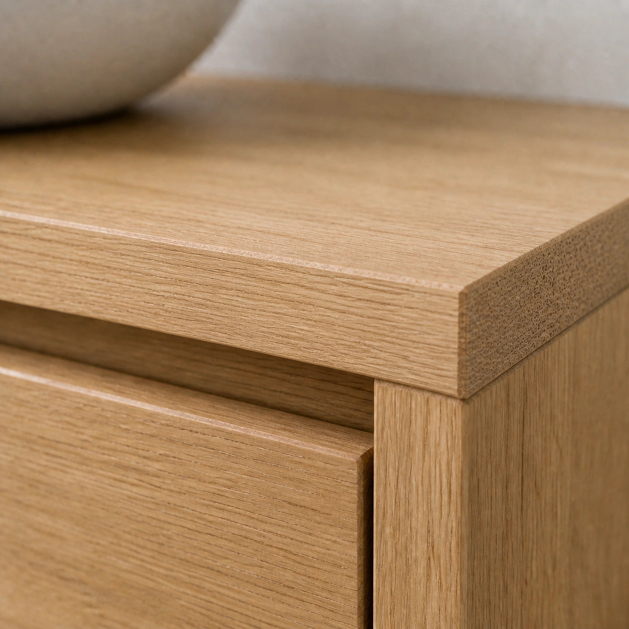 Close-up of a light oak wood furniture piece, showing the drawer front, top edge, and side corner with a neutral textured background.