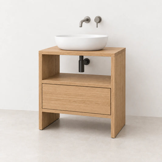 A modern single-drawer bathroom vanity cabinet in light oak wood, featuring an open shelf, a white vessel sink, and a matte black drain, set against a minimalist off-white wall and light concrete floor.